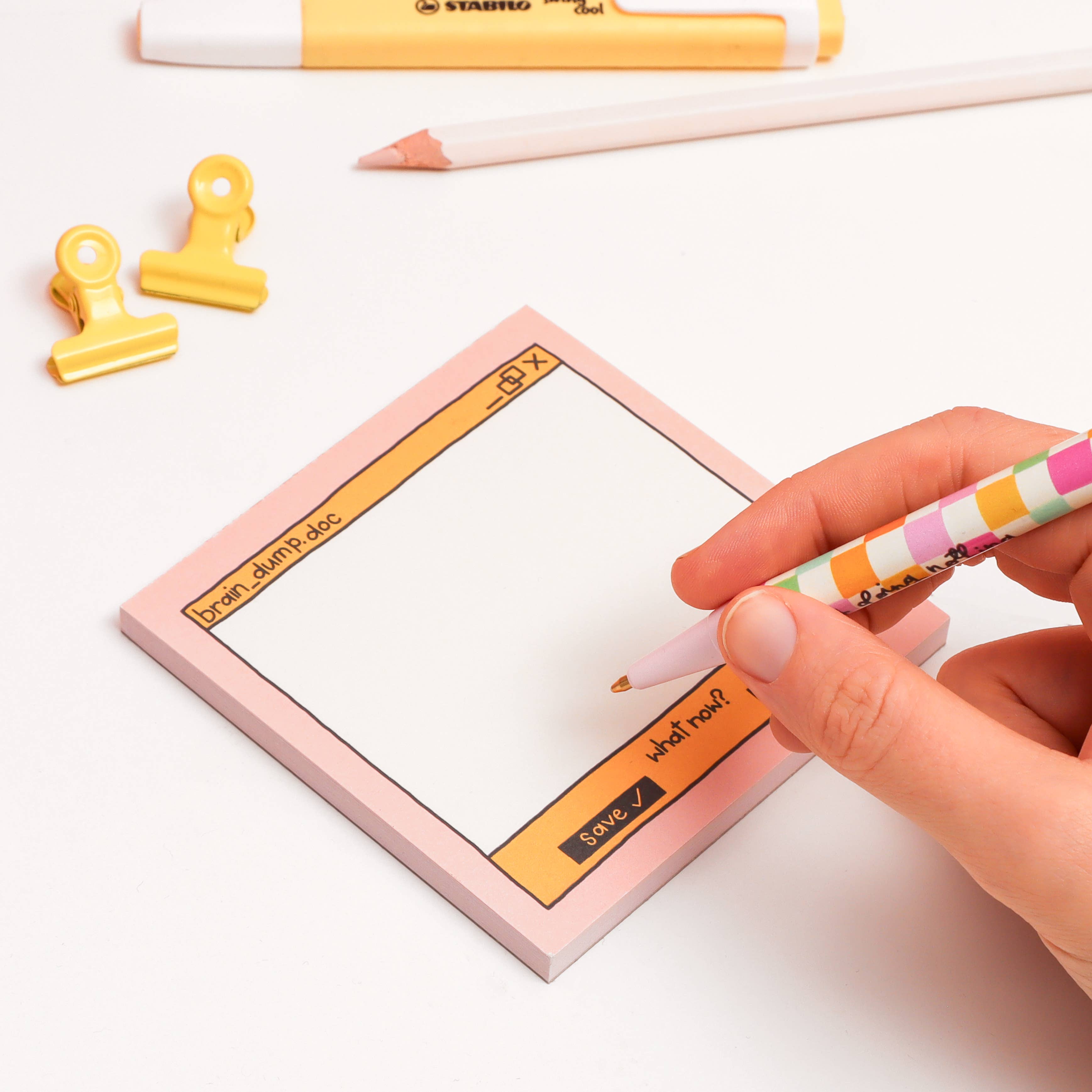 Person writing on a pink notepad with a colorful pen, surrounded by office supplies on a white background.