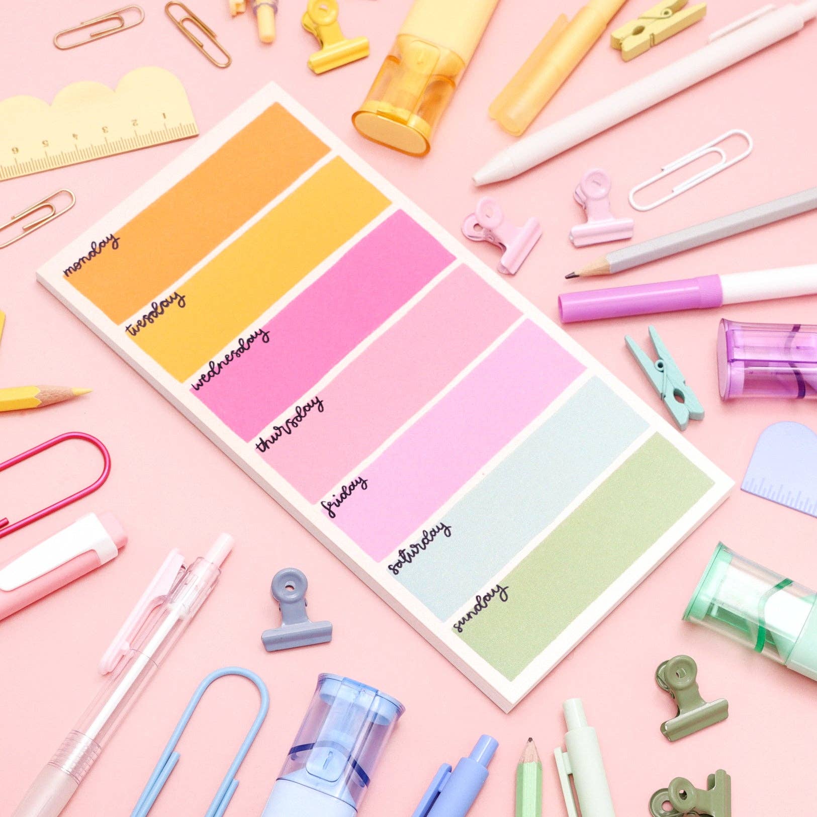 Colorful note pad with days of the week on a pink background with stationery items.